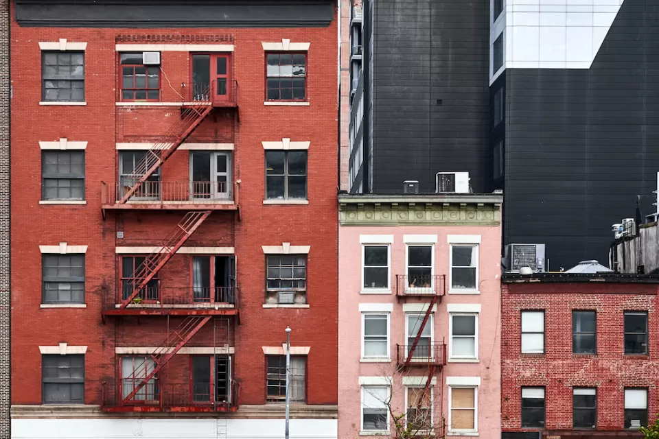 Three brick apartment buildings of various heights with fire escapes in New York City.